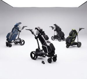A studio comparison shot featuring four different motorized electric golf trolleys with loaded bags (blue, black/white, all-black, and grey/lime) arranged on a seamless white background, including the central MGI Zip Navigator with its remote.